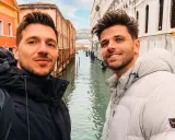 Two men enjoying a romantic gay Venice trip, smiling for a selfie with canals and historic buildings behind them.
