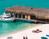A boat docks at a thatched-roof beach bar on a sunny day. This scene embodies the welcoming spirit of Aruba, a destination known for being gay friendly.