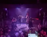 Performers in masks and elaborate outfits dance on stage at one of Valencia's vibrant gay bars, while a crowd watches below.