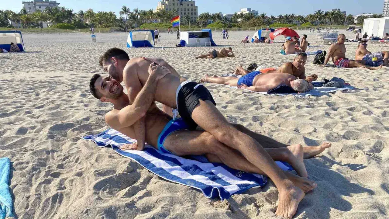 Two men embrace on a towel at Haulover Beach, a popular spot for gay beachgoers. Other sunbathers relax on the sand in the background.