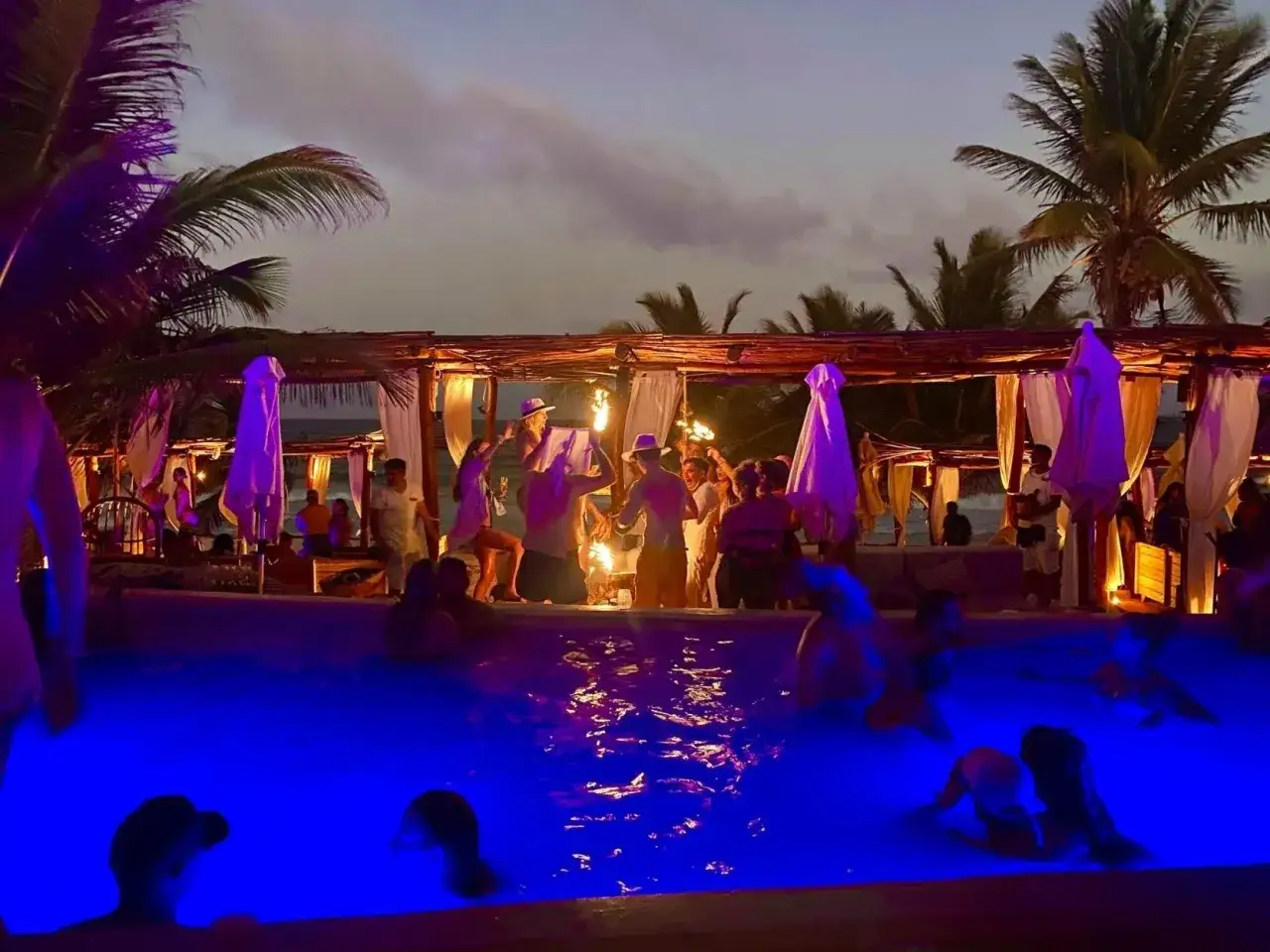 A vibrant gay Tulum party unfolds at dusk. People dance by torches near a pool lit with blue light, with palm trees silhouetted against the sky.