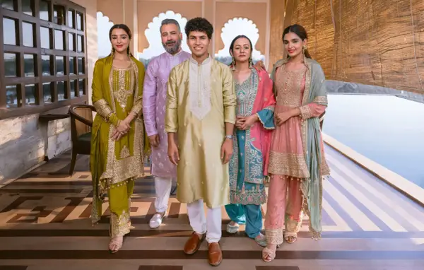 A family poses in vibrant traditional clothing from India. The women wear ornate salwar kameez sets in olive green, teal, and pink, while the men are dressed in elegant kurtas.