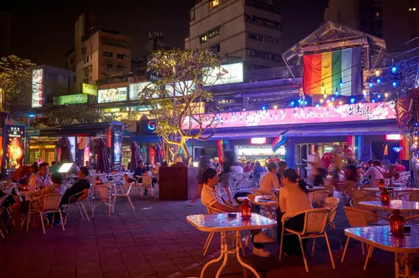 A vibrant night scene in gay Taipei, with people dining outdoors under colorful lights and a rainbow flag.