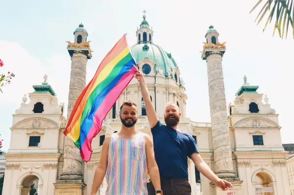 Two men proudly hold a rainbow flag in front of a grand building in Austria, celebrating gay pride.