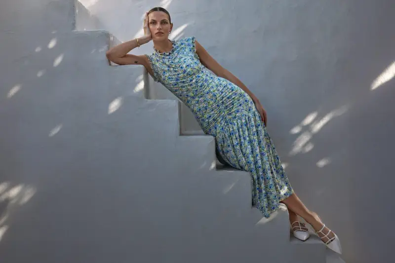 A woman in a floral dress and white heels, embodying a chic formal dress code for women, poses on white stairs.